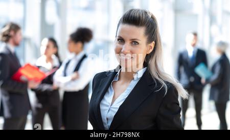 Schöne junge Geschäftsfrau in formellem Anzug, die vor Kollegen im Geschäftsgebäude steht Stockfoto