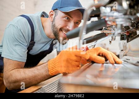 Fröhlicher junger Mann, der die Kaffeemaschine im Café repariert Stockfoto