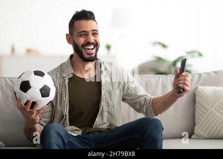 Glücklicher junger arabischer Mann, der Fußballspiel im Fernsehen sieht, den Ball und den Controller zu Hause hält Stockfoto