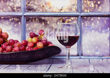 Ein paar lila Trauben, ein Glas Rotwein oder Saft stehen auf einem Holztisch gegen das Fenster. Rote Traube auf einem Teller. Ernte im Weinberg. Romantischer Abend Stockfoto