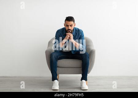 Arabischer Mann, der im Sessel sitzt, mit dumpfem Gesichtsausdruck, sich traurig oder müde fühlt, Probleme hat, an Depressionen leidet Stockfoto