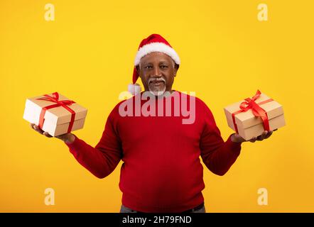 Älterer schwarzer Mann im Weihnachtsmannhut hält Weihnachtsgeschenke in beiden Händen über orangenen Studiohintergrund Stockfoto