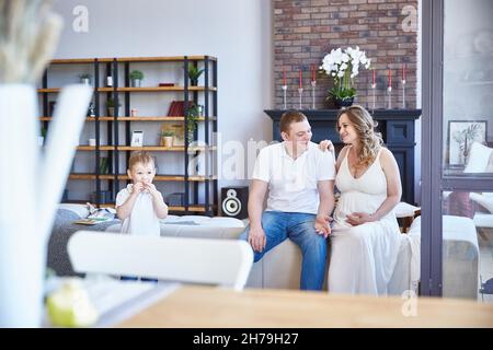 Eine glückliche Familie mit einem kleinen Kind und einer Schwangeren sitzt auf dem Sofa im Wohnzimmer. Stockfoto