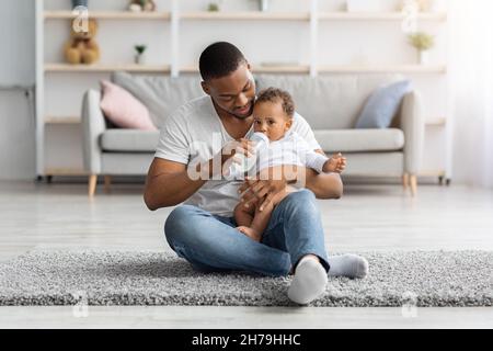 Konzept Für Die Kinderbetreuung. Liebevoller Afroamerikanischer Vater, Der Baby Flasche Mit Wasser Gibt Stockfoto