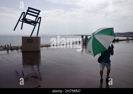 Mann & Regenschirm am Regentag auf der Promenade des Anglais mit Big Blue Chair Skulptur von Sabine Geraudie Nice Alpes-Maritimes Frankreich Stockfoto