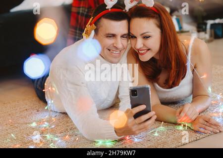 Ein glückliches Ehepaar liegt zu Hause in der Nähe des Weihnachtsbaums auf dem Boden und fotografiert sich selbst. Stockfoto