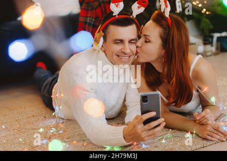 Ein glückliches Ehepaar liegt zu Hause in der Nähe des Weihnachtsbaums auf dem Boden und fotografiert sich selbst. Stockfoto