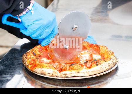 Die Hände des Kellners in blauen Latexhandschuhen schneiden die gebackene Pizza mit einem runden Metallmesser. Speicherplatz kopieren. Stockfoto