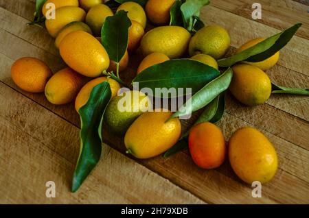 Kleine tropische Zitrusorangen von Kumquat mit dunkelgrünen länglichen glänzenden Blättern liegen auf einem Holzbrett. Stockfoto