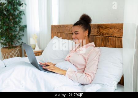 Aufgeregte afroamerikanische Frau im Schlafanzug, die unter der Decke auf dem Bett sitzt und mit einem Laptop unterwegs von zu Hause aus arbeitet Stockfoto