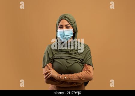 Coronavirus-Konzept. Junge muslimische Frau in Hijab trägt Schutzmaske und posiert mit gekreuzten Händen Stockfoto