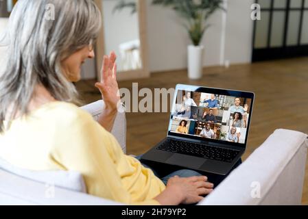 Moderne Ruhestand Frau begrüßt Teilnehmer von Online-Meeting, hat Videokonferenz mit Kollegen auf der Ferne, ältere Frau auf dem Sofa liegend, mit Laptop für Videoanruf an diverse Team Stockfoto