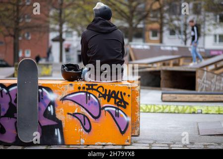 Junger Mann, der am Rande eines Skateparks sitzt und sein Skateboard dabei betrachtet, wie seine Kollegen Tricks vorführen Stockfoto