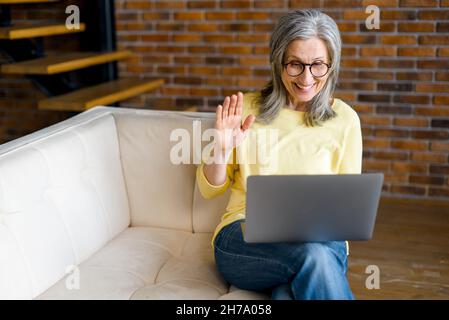 Eine glückliche, ältere, grauhaarige Geschäftsfrau im mittleren Alter, die auf der Couch sitzt, die Webcam anschaut und winkt, Studenten, Teilnehmer einer Online-Konferenz begrüßt und von zu Hause aus ein virtuelles Meeting auf dem Laptop führt Stockfoto