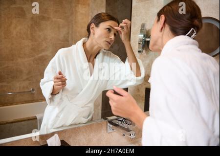 Schöne europäische Brünette mittleren Alters Frau trägt weißen Frottee Bademantel Blick auf ihre Reflexion im Badezimmerspiegel während der morgendlichen Hygiene rou Stockfoto