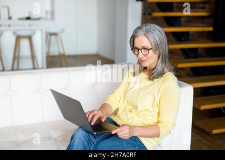 Fokussierte, reife Frau in einer stilvollen Brille mit einem Laptop, die von zu Hause aus in entspannter Atmosphäre arbeitet, charmante ältere Frauen konzentrierten sich auf die Tastatur, die auf der Couch sitzt Stockfoto