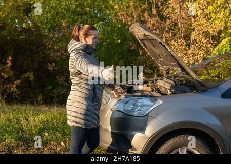 Eine verärgerte Frau steht in der Nähe ihres kaputten Autos und spricht im Herbst auf einem Handy auf der Straße. Stockfoto