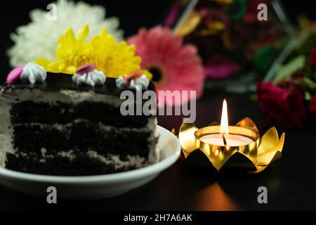 Goldene Teelichtkerze In Lotus-Förmigem Halter Mit Einem Stück Schokoladentrüffelkuchen An Der Seite Und Bunten Frischen Blumen Auf Dunklem Schwarzem Backgr Stockfoto