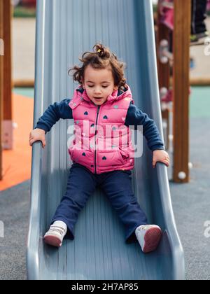 Kleines Mädchen, das Spaß hat, die Rutsche hinunter zu rutschen, während es auf dem Spielplatz im Park spielt. Stockfoto