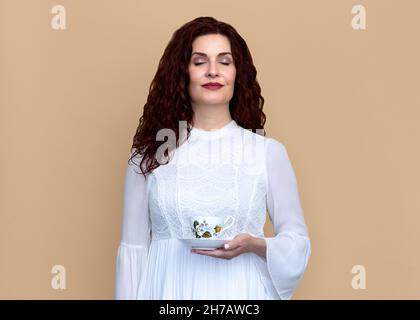 Frau mit geschlossenen Augen hält und riecht Tee mit Beige Hintergrund. Sie trägt ein romantisches weißes Kleid und hat welliges rotbraunes Haar und ein weiches Lächeln. Stockfoto