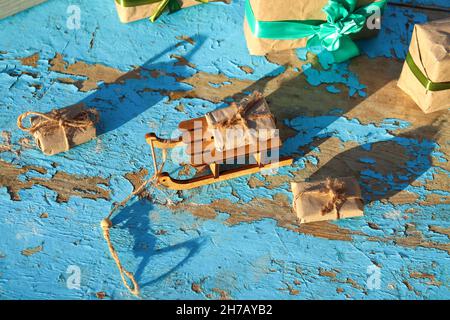 Geschenk. Gewickelt Handwerk vintage Geschenkbox liegen Holzspielzeug Schlitten auf blauem alten Holzhintergrund. Geschenk-Box in Bastelpapier. Urlaubskonzept. Weihnachten, Weihnachten Stockfoto