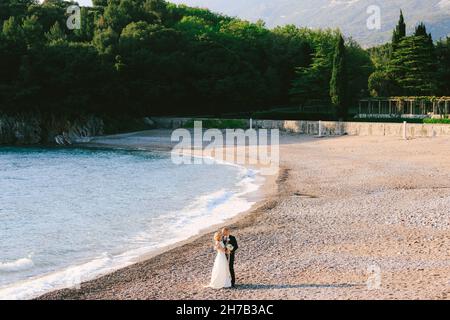 Bräutigam in einem blauen Anzug umarmt und küsst Braut in einem weißen Kleid mit einem Blumenstrauß, der am Strand in der Nähe des Meeres vor dem Hintergrund steht Stockfoto