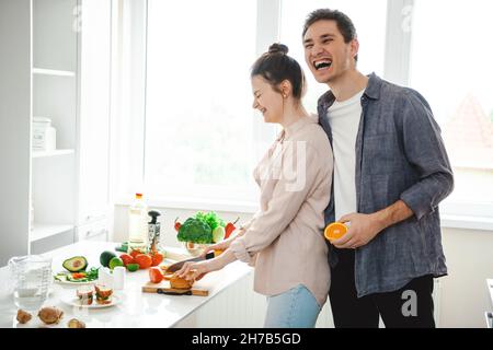 Mann und Frau schneiden Gemüse, umarmen, reden, lachen, Spaß beim Kochen zusammen gesunde Mahlzeit in der Küche vorzubereiten. Lächelnder Mann. Stockfoto