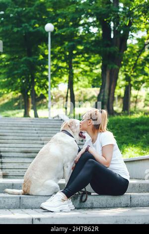 Porträt einer Frau, die mit ihrem Hund labrador auf der Treppe sitzt, küsst und umarmt. Morgenspaziergänge. Freundschaft zwischen Mensch und Hund Stockfoto