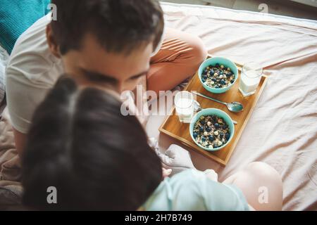 Blick von oben auf ein Paar, das sich küsst, während es ein gesundes Frühstück im Bett hat. Gesunder Lebensstil. Gesunde Ernährung. Stockfoto