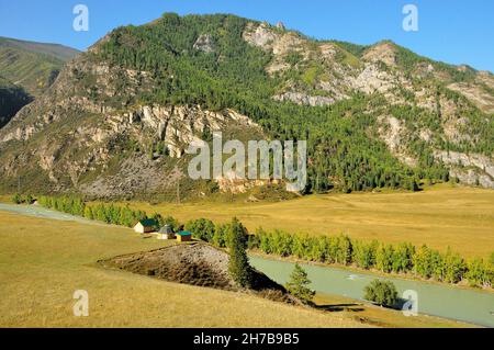 Mehrere kleine Häuser stehen am Rande eines hohen Hügels am Fluss, der durch das malerische Tal am Fuße der felsigen Berge fließt. Katun Ri Stockfoto