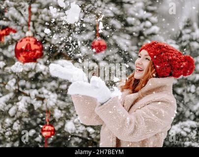 Junge lächelnde schöne Frau in Pelzmantel und Fäustlingen in der Nähe geschmückten Weihnachtsbaum stehen und Schneeflocken im Freien fangen Stockfoto