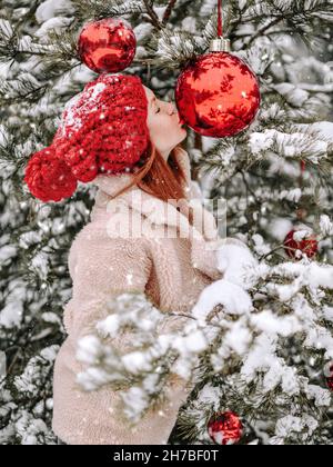 Junge Frau im Pelzmantel steht draußen am geschmückten Weihnachtsbaum und küsst einen großen roten Ball aus Neujahrsfichte unter Schnee Stockfoto