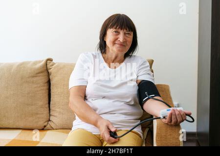 Erwachsene weibliche Person, die ihren Blutdruck und Puls mit einem elektronischen Gerät misst. Reife Frau zu Hause auf dem Sofa Stockfoto