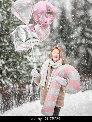 Junge lächelnde schöne Frau in stilvollem Pelzmantel und Fäustlingen zu Fuß mit Spielzeug Zauberstab und Luftballons bei Schneefall im Freien Stockfoto