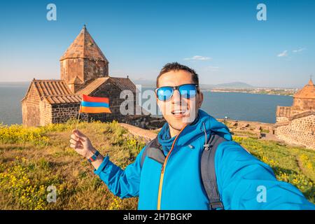 Ein Mann mit armenischer Flagge macht ein Selfie-Foto vor dem Hintergrund des Klosters Sevanavank am Sevan-See. Urlaub und Patriotismus Konzept Stockfoto