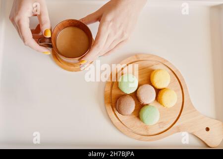 Frühstück am Morgen auf weißem Tisch. Tasse Kaffee mit Milch in den Händen der Frau und Holzschale mit Makronen. Süßes Dessert und heißes Getränk. Stockfoto