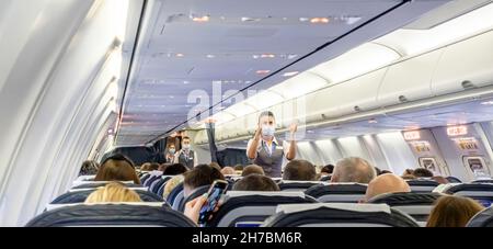 Weibliche Flugbegleiterin in Gesichtsmaske, die Passagieren, die in einem Flugzeug der Boeing 737-800 Belavia auf Stühlen sitzen, Notausgänge zeigt Stockfoto