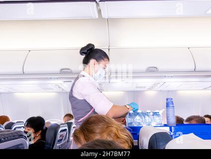 Eine weibliche Flugbegleiterin in Gesichtsmaske serviert den Passagieren, die in Stühlen in einer Flugzeugkabine eines Belavia Boeing 737-800-Flugzeugs sitzen, Wassergetränke Stockfoto
