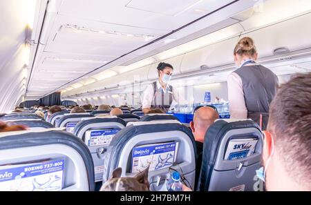 Weibliche Flugbegleiter in Gesichtsmasken servieren Getränke an Passagiere, die in Stühlen in einer Flugzeugkabine eines Belavia Boeing 737-800-Flugzeugs sitzen Stockfoto