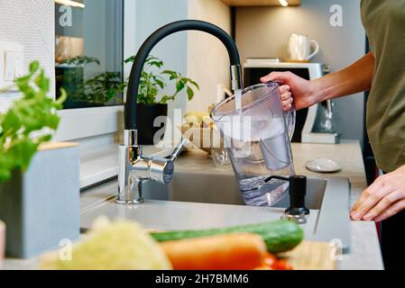 Frau goss Wasser aus dem Wasserhahn in den Wasserfilterkrug in der Küche Stockfoto