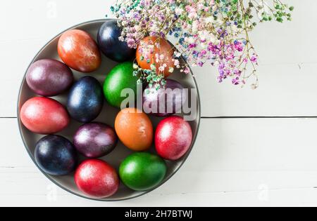 Ein großes Gericht mit bunten Ostereiern und Blumen auf weißem Holzhintergrund. Draufsicht. Traditionelle Osterleckereien Stockfoto