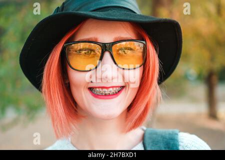 Ginger Mädchen mit Zahnspangen und Piercings in gelben Gläsern und stilvollen Hut lächelt Blick auf die Kamera. Junge Frau mit kieferorthopädischer Halterung. Richtiger Biss c Stockfoto