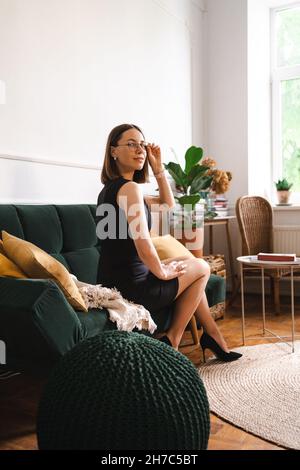 Selbstbewusste Geschäftsfrau mit Brille in einem schwarzen klassischen Kleid, die in einem modernen Apartment auf einer Couch sitzt. Grünes Plüschsofa mit gelben Kissen Stockfoto