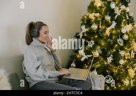 Fröhliche kaukasische Frau, die am Weihnachtstag in der Nähe des Weihnachtsbaums Musik hört. Attraktive Frau sitzt auf der Couch genießen Sie Songs in Kopfhörer und Laptop. Stockfoto