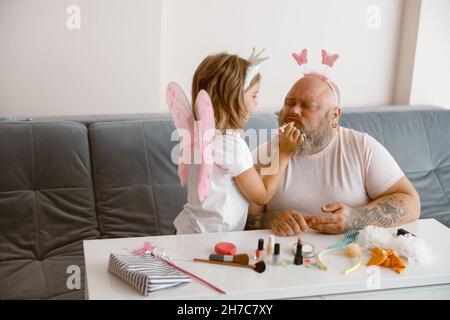 Kleine Tochter mit Flügeln trägt im Wohnzimmer Lippenstift auf den Mund des Vaters auf Stockfoto