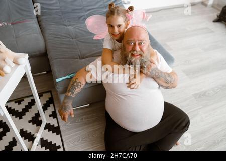 Glückliches kleines Mädchen umarmt lustig übergewichtigen Dadipanten sitzen auf dem Boden im Wohnzimmer Stockfoto