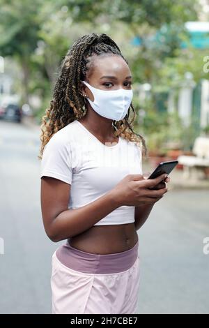 Schöne Frau mit Smartphone im Freien stehend, SMS an Freundin und Blick auf die Kamera Stockfoto