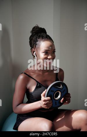 Smilng ziemlich fit junge Frau sitzt auf Fitness-Ball und macht Übungen mit Hantelscheibe Stockfoto