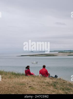 Ein Touristenpaar blickt auf eine Segelyacht in der Küstenlandschaft von Holy Island Lindisfarne in Northumberland England - Nordseeküste Stockfoto