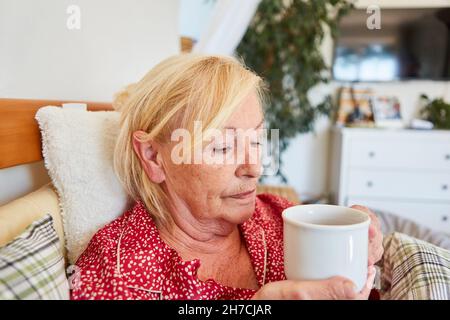 Die alte Frau liegt traurig und nachdenklich im Bett zu Hause und trinkt eine Tasse Kaffee Stockfoto
